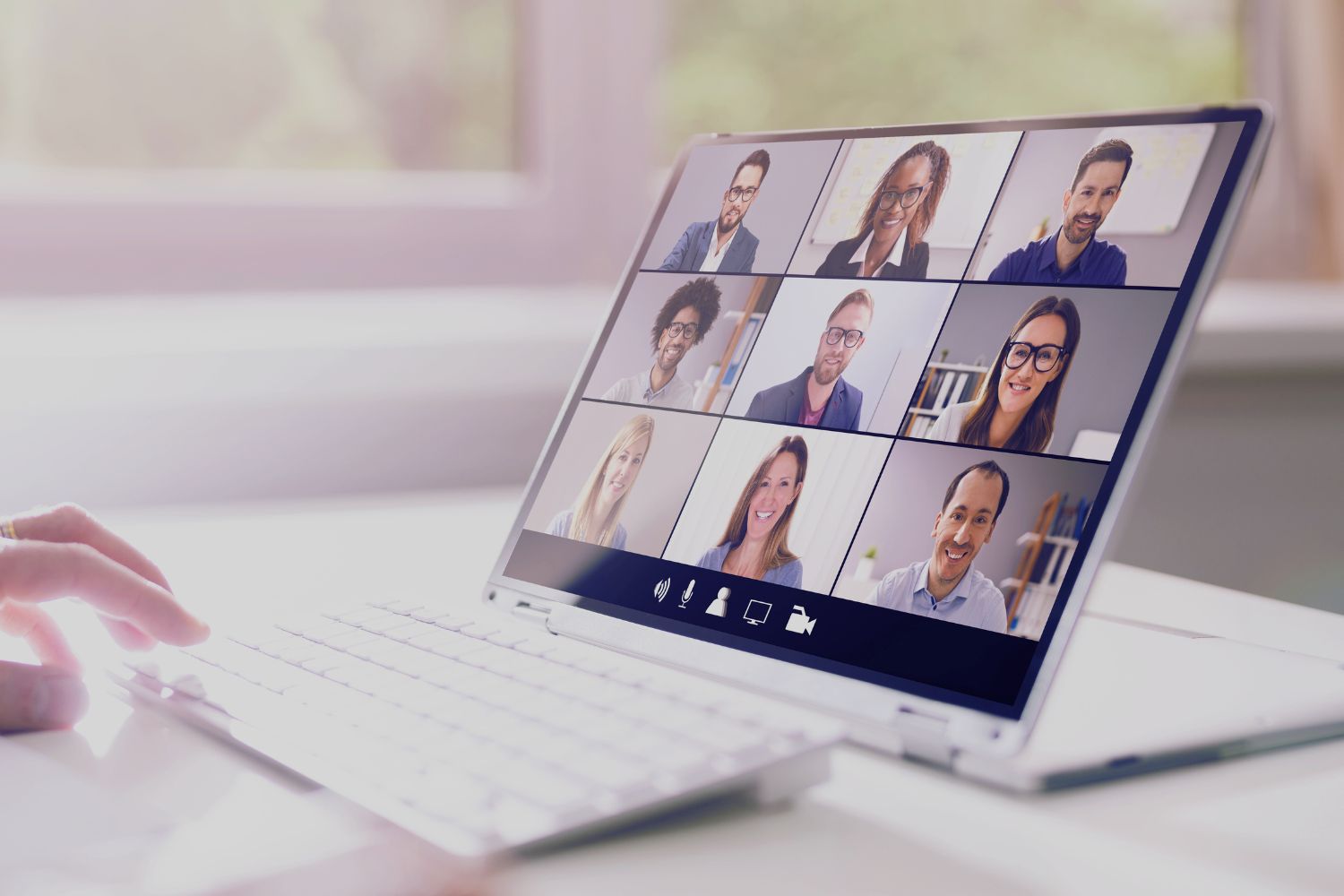 Picture of a zoom meeting with 9 people on a screen on top of a desk next to a window